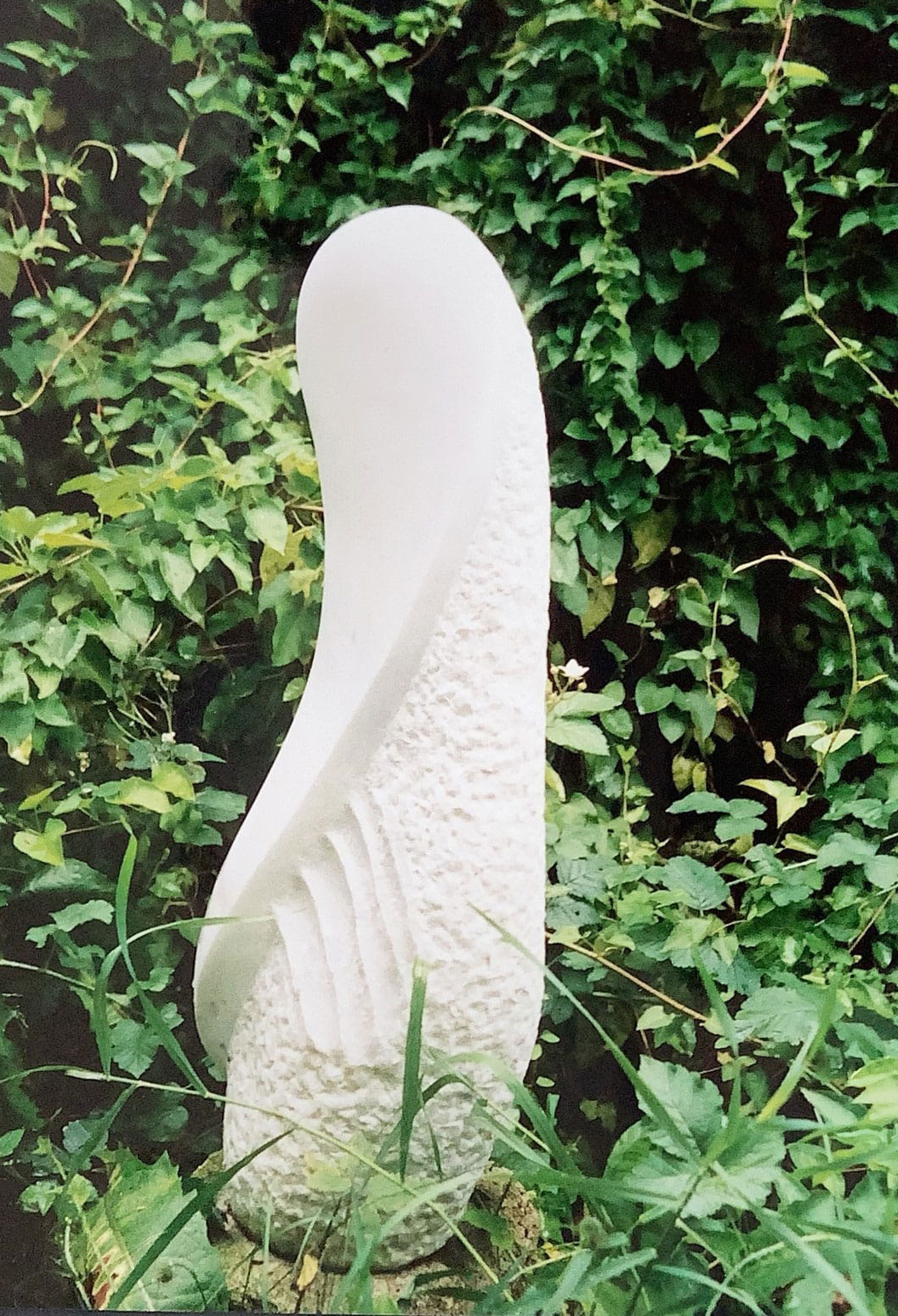 Eine schlanke Marmorskulptur, die eine gebogene Linie zeigt, steht in einem Garten.