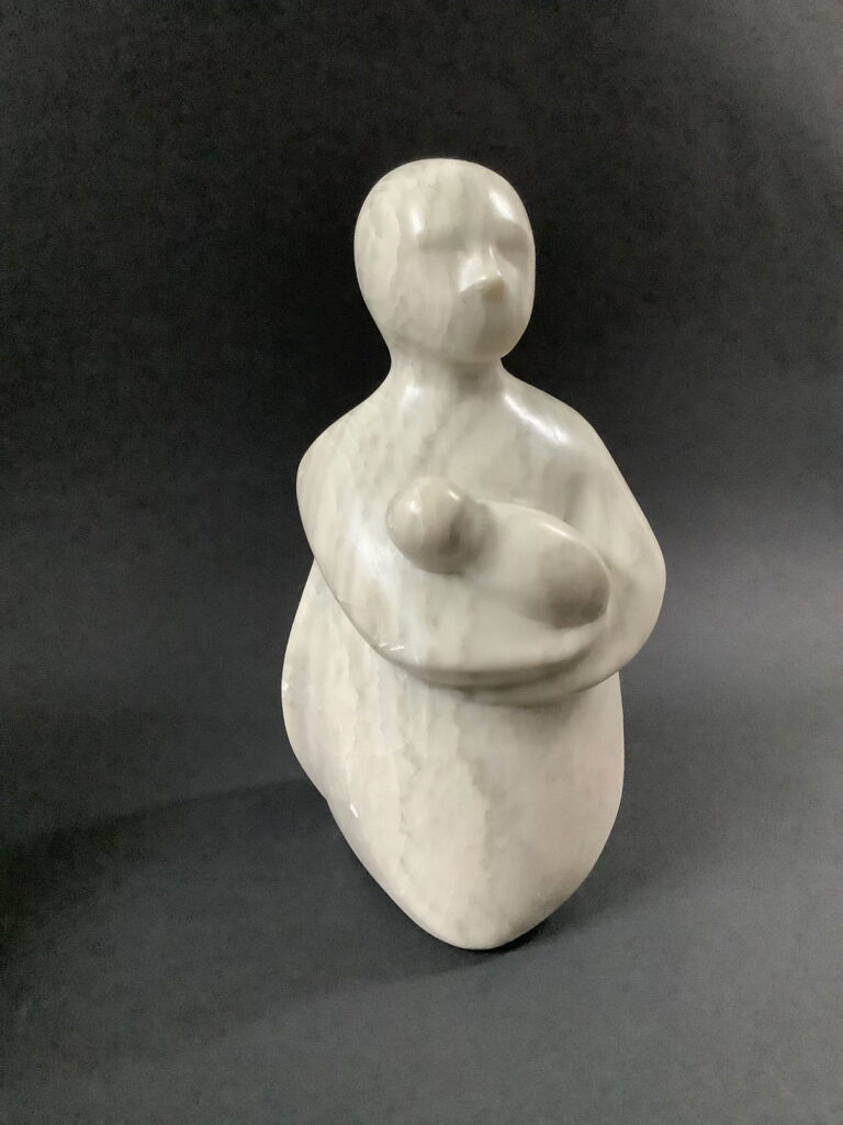 Kleine Skulptur aus grauem Speckstein von der Seite gesehen.. Eine stark abstrahierte Frauenfigur trägt ein Baby im Arm.