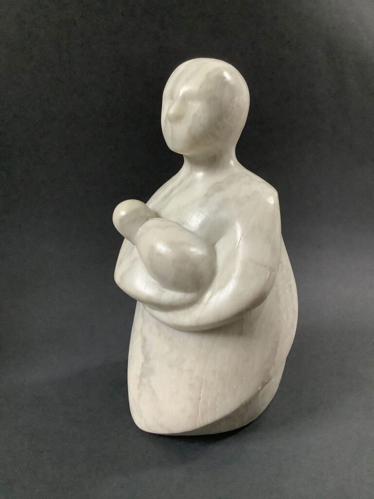 Kleine Skulptur aus grauem Speckstein von schräg vorne gesehen.. Eine stark abstrahierte Frauenfigur trägt ein Baby im Arm.