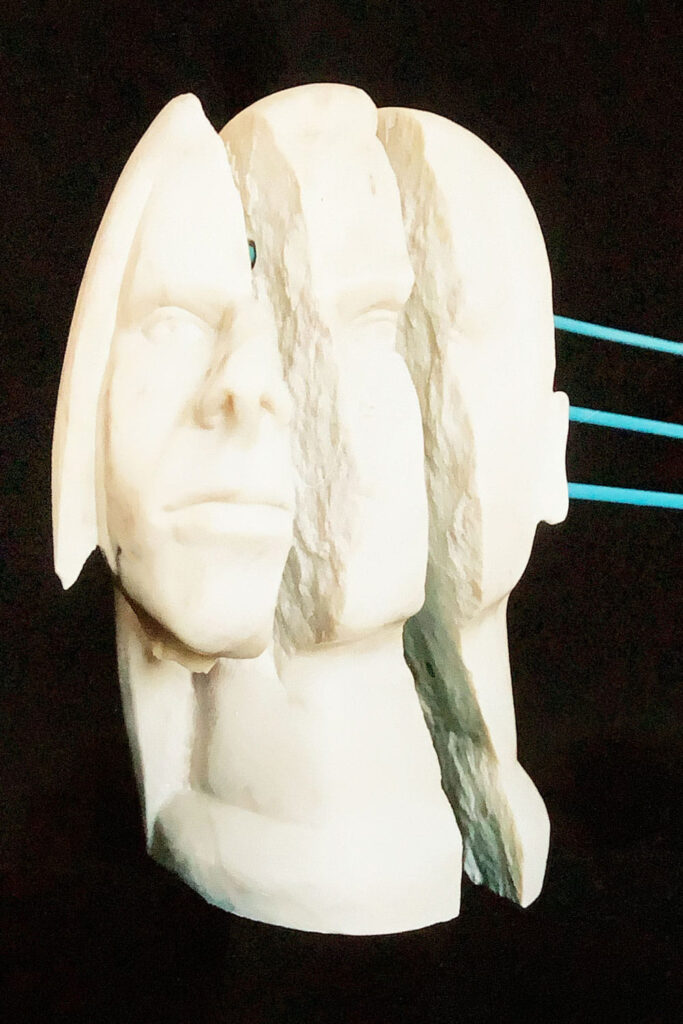 Skulptur aus weissem Speckstein. Kopf eines jungen Mannes von vorne, der Stein ist zerteilt, aber mit 3 blauen Metallstäben verbunden